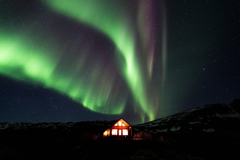 Sweeping green aurora above a glowing red wooden cabin near Snæfellsnes in Iceland at night.