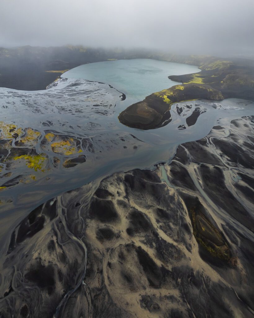 Aerial of a teal Highland lake feeding braided silt rivers across black sand under low cloud, Iceland.