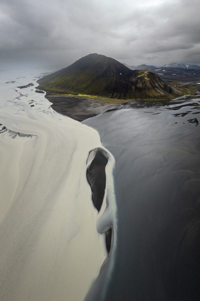 Aerial of a moss-green peak above a wide black-sand plain with snow-streaked channels, Icelandic Highlands.