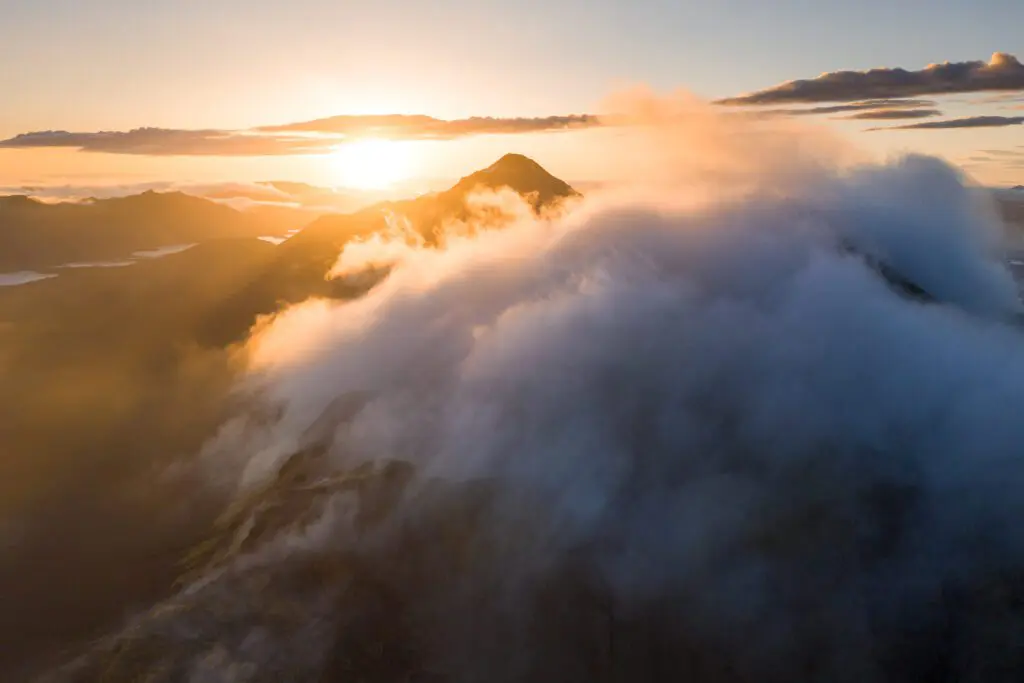 Aerial of a sharp peak wreathed in cloud above pastel Icelandic lake landscape.