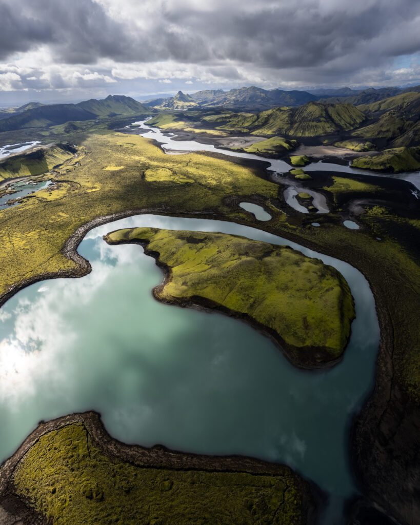 Wide aerial of moss-green Highland hills curving around a teal lake under storm clouds, Iceland.