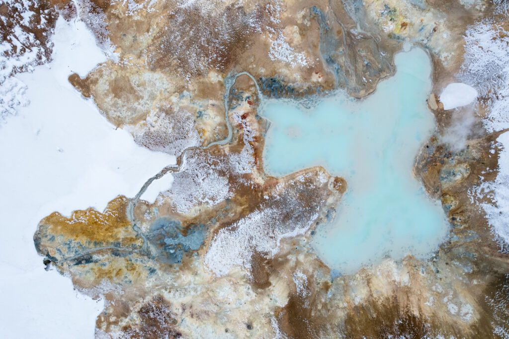 Top-down aerial of a pale-blue geothermal pool in red-orange mineral mud, Iceland.