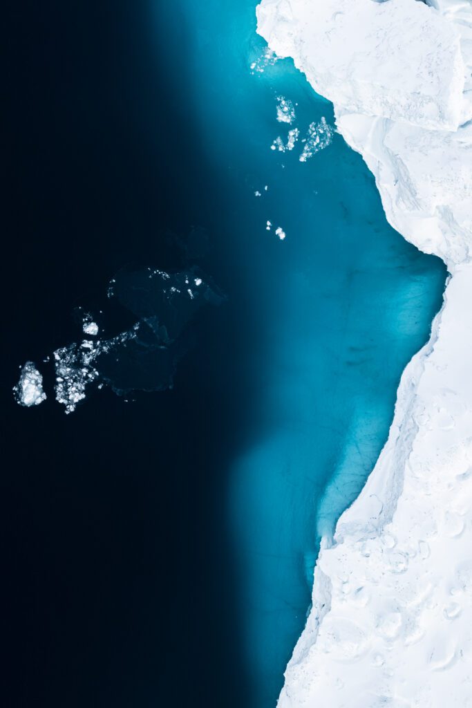 Top-down drone view of a small ice floe drifting beside a sharp iceberg edge in turquoise water, Greenland.