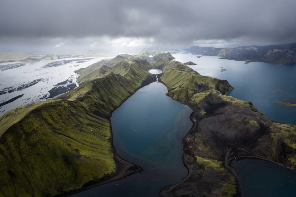Aerial of green Highland ridges curving around a deep teal lake under storm clouds, Iceland.