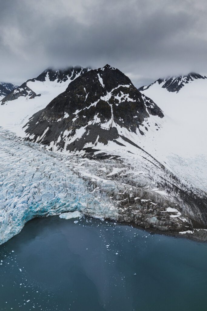 Aerial of a sharp dark peak above a crevassed blue glacier face meeting still fjord water.
