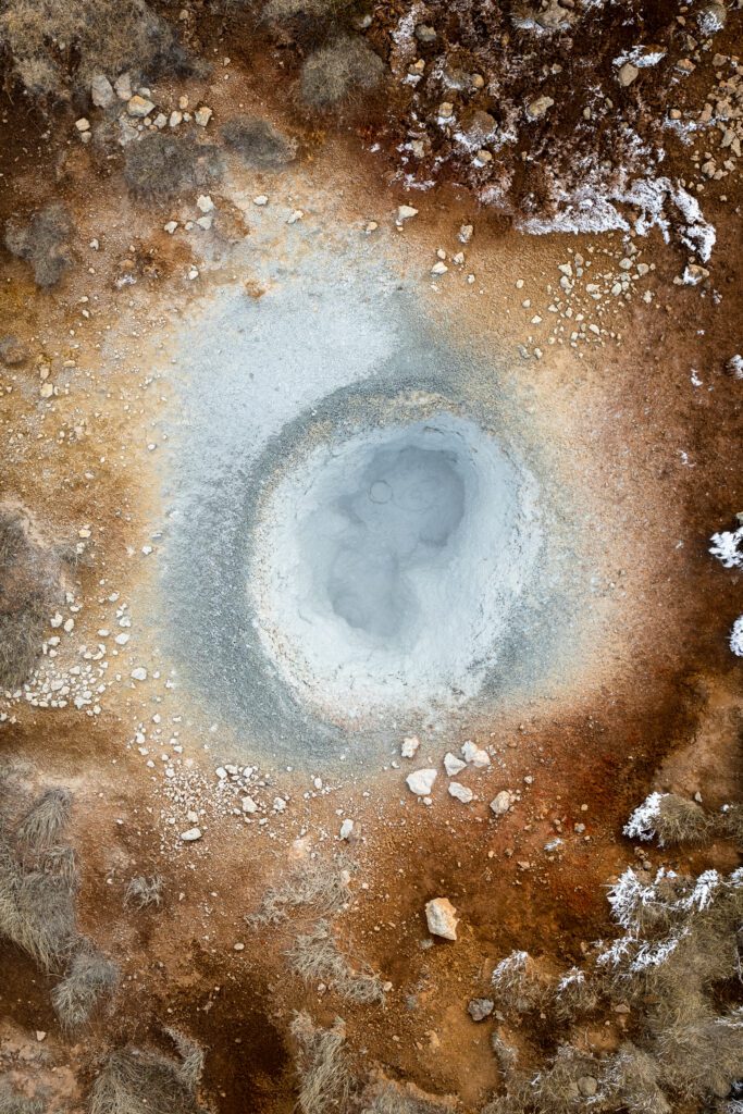 Top-down aerial of a steaming geothermal mud pot ringed by rust-coloured earth, Iceland.