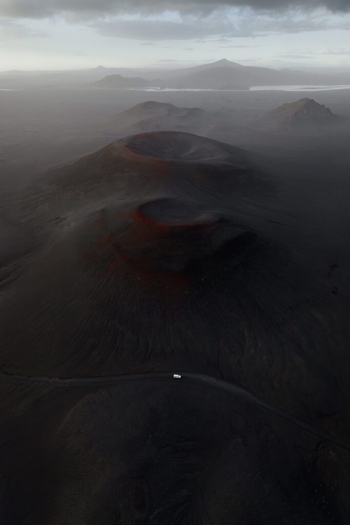Aerial of two black cinder cones with red rims in foggy lava fields, distant peaks behind, Iceland.