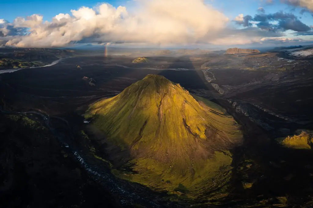 Aerial of a sunlit green pyramid peak with a faint rainbow beyond, Icelandic Highlands.