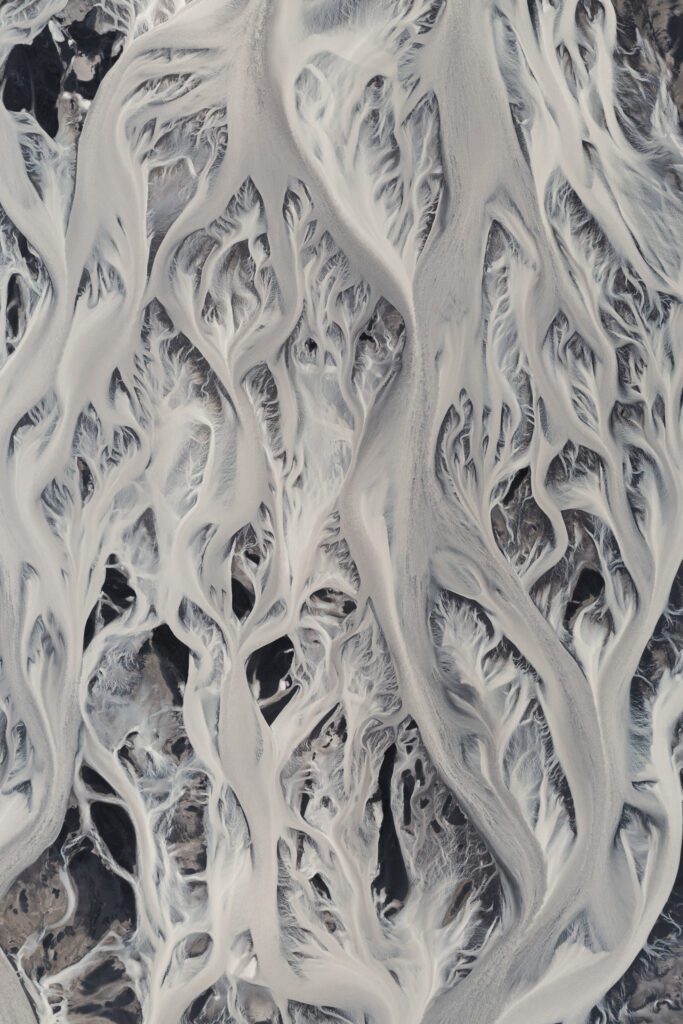 Abstract aerial of pale braided river channels weaving through dark Icelandic plains.