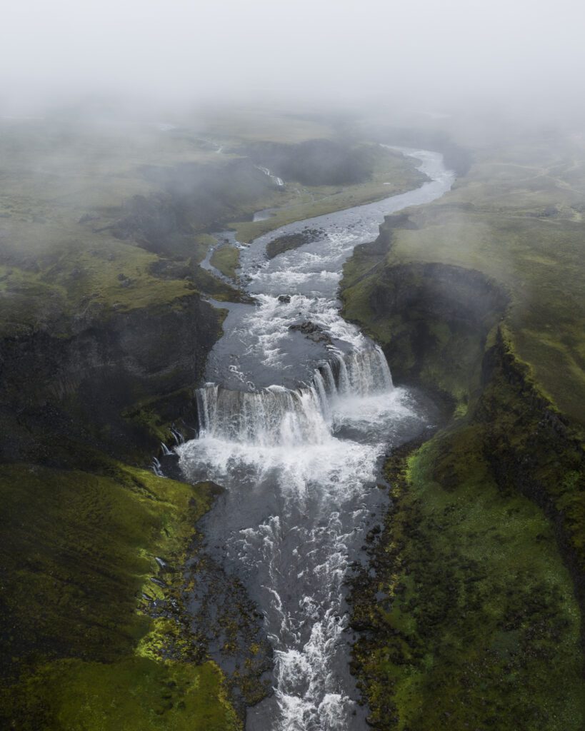 Aerial of a roaring waterfall stepping down a misty green Highland canyon, Iceland.