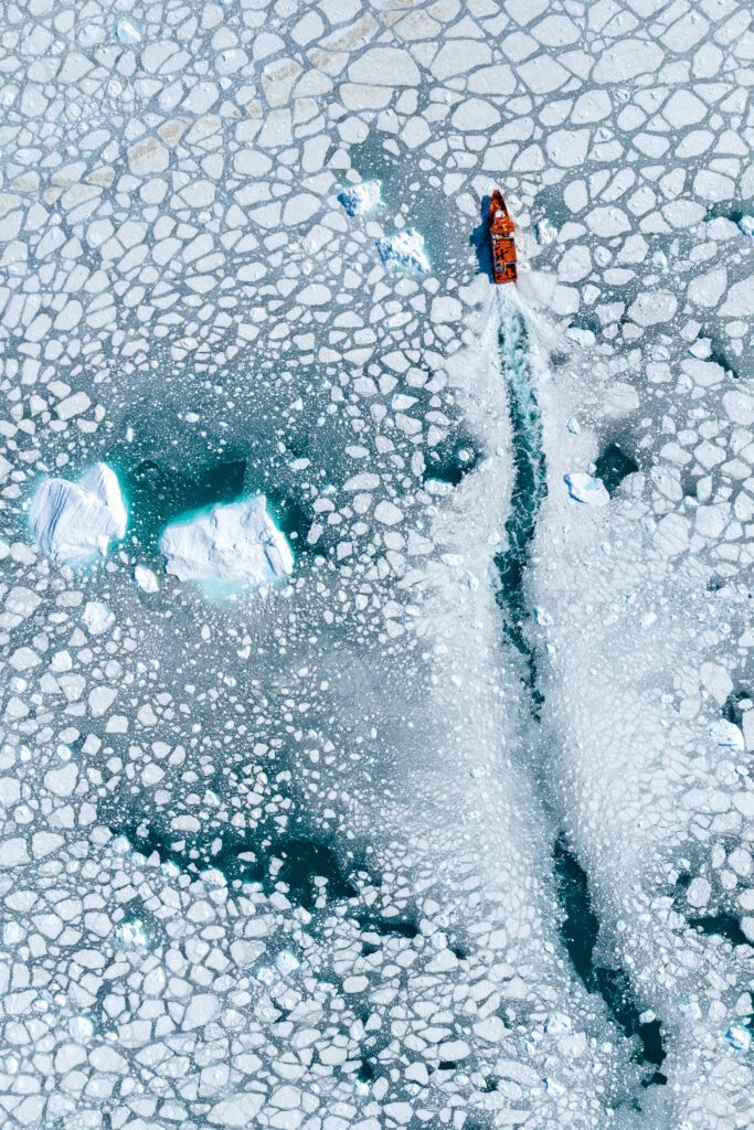 Top-down aerial of a red boat threading through pancake ice past an iceberg in Greenland.