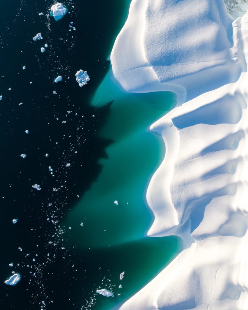 Top-down aerial of turquoise meltwater along the sculpted edge of a Greenland iceberg.