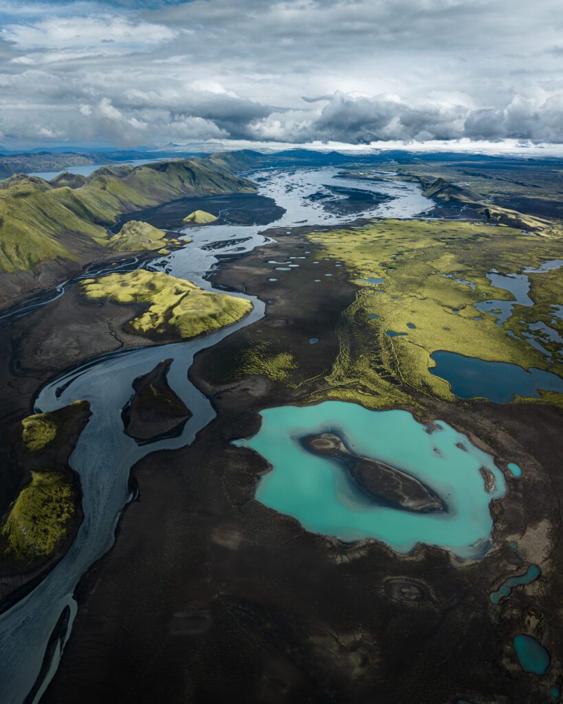 Wide aerial of a turquoise meltwater pool among dark sand braids and mossy Highland patches, Iceland.