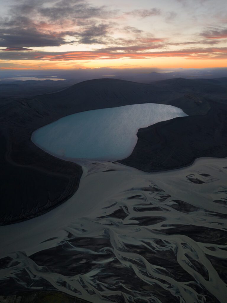 Sunset aerial of a pale-blue lake cradled in dark hills above black-sand river braids, Iceland.