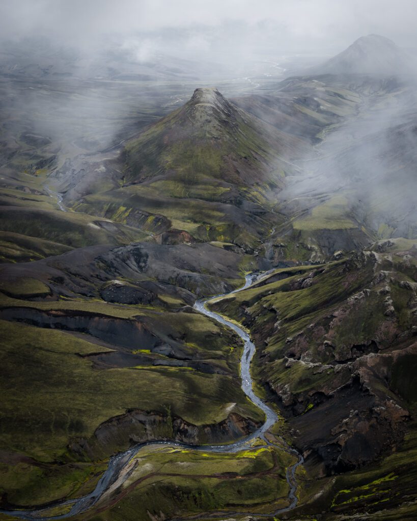 Aerial of a misty green Highland canyon with a river winding past a pyramid peak, Iceland.