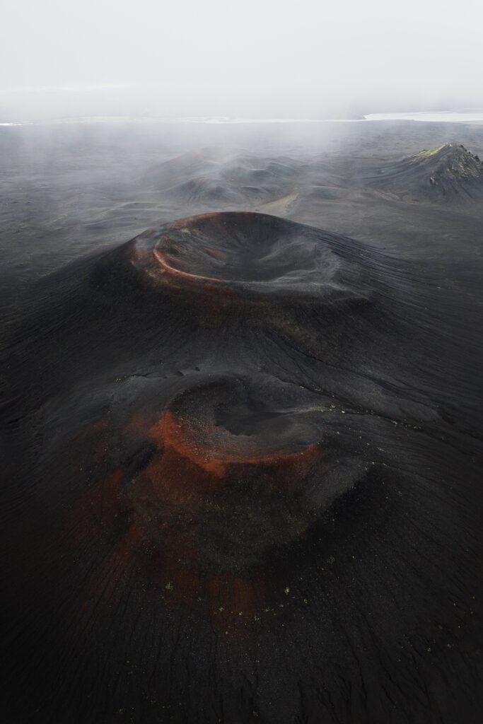 Aerial of two red-rimmed cinder cones rising from a foggy black volcanic plain, Iceland.