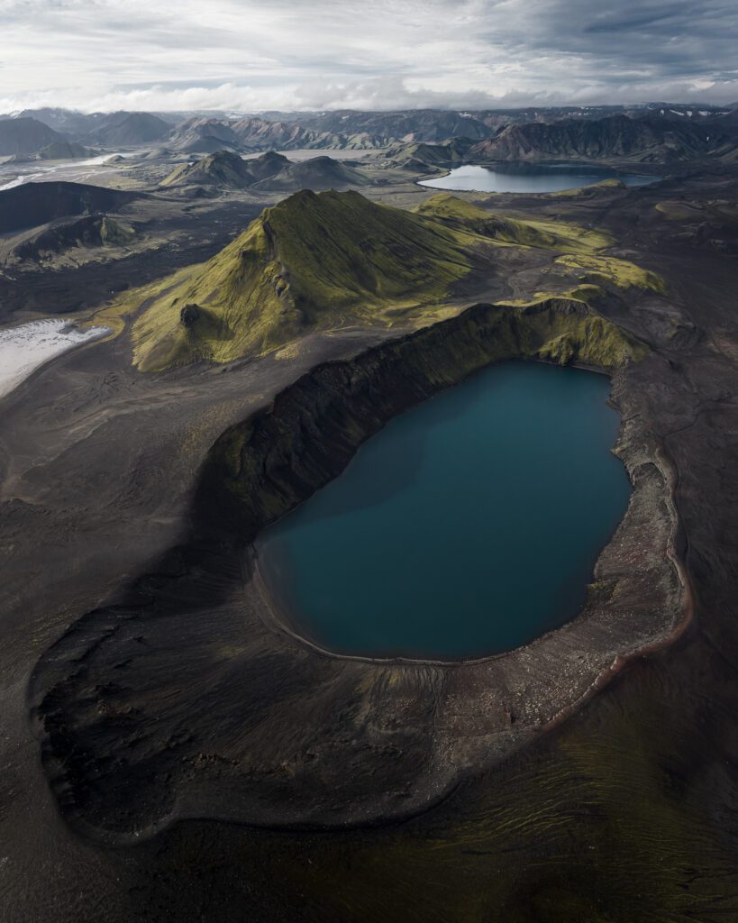 Aerial of the deep teal Bláhylur crater lake in its mossy black-sand rim, Icelandic Highlands.