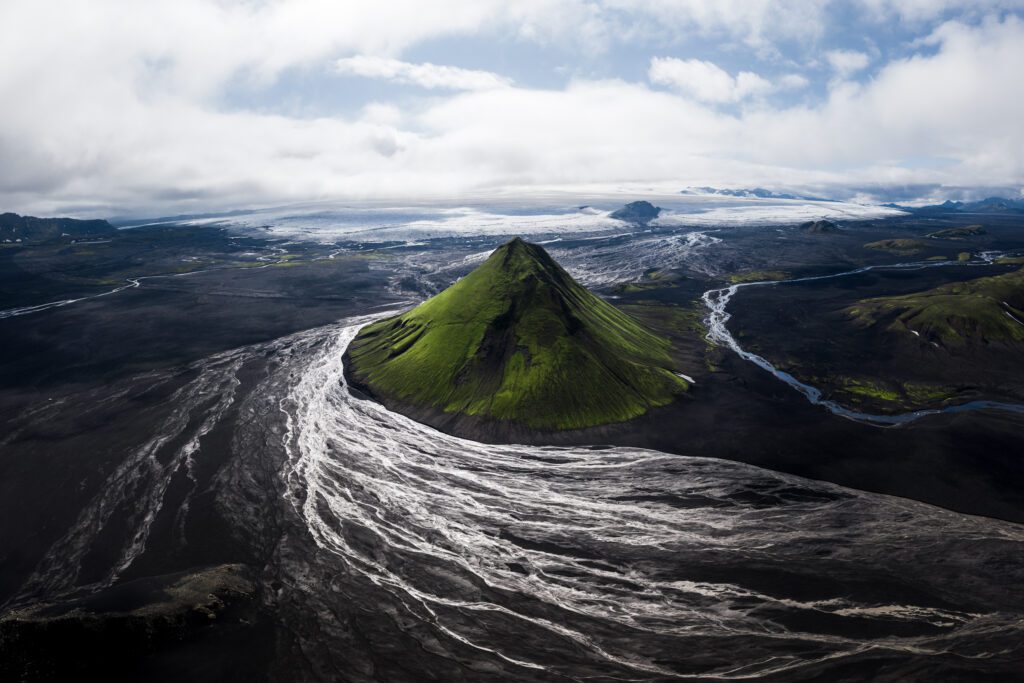 Aerial of the green pyramid of Mælifell rising from a wide black-sand braided plain, Iceland.