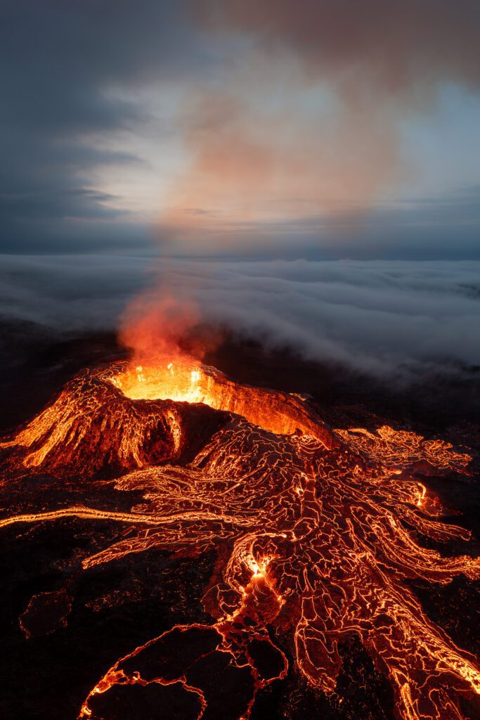 Aerial drone shot of a volcano erupting through low clouds with lava flowing downslope in Iceland.