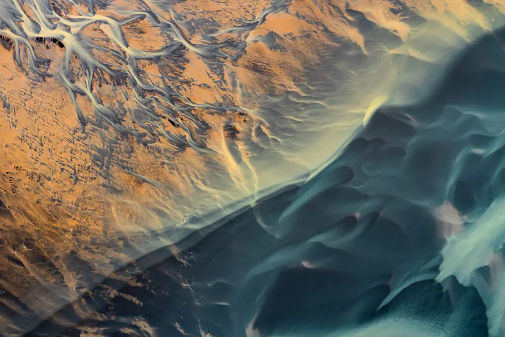 Abstract aerial of sunlit braided river channels meeting smooth dark sand in the Icelandic Highlands.