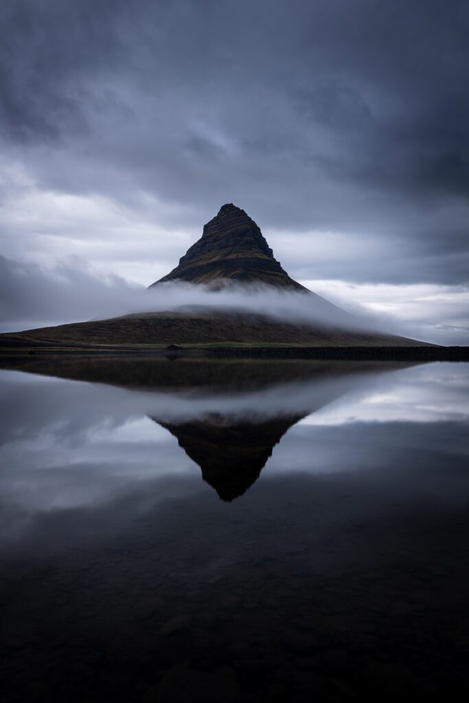 Kirkjufell reflected in glass-still water under heavy overcast at twilight.