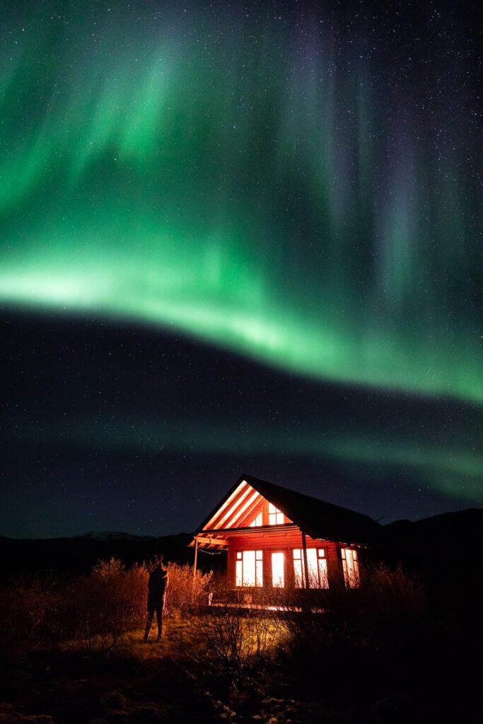 Green aurora arching above a red-lit cabin in Hraundalur with a tiny figure watching from the lawn.