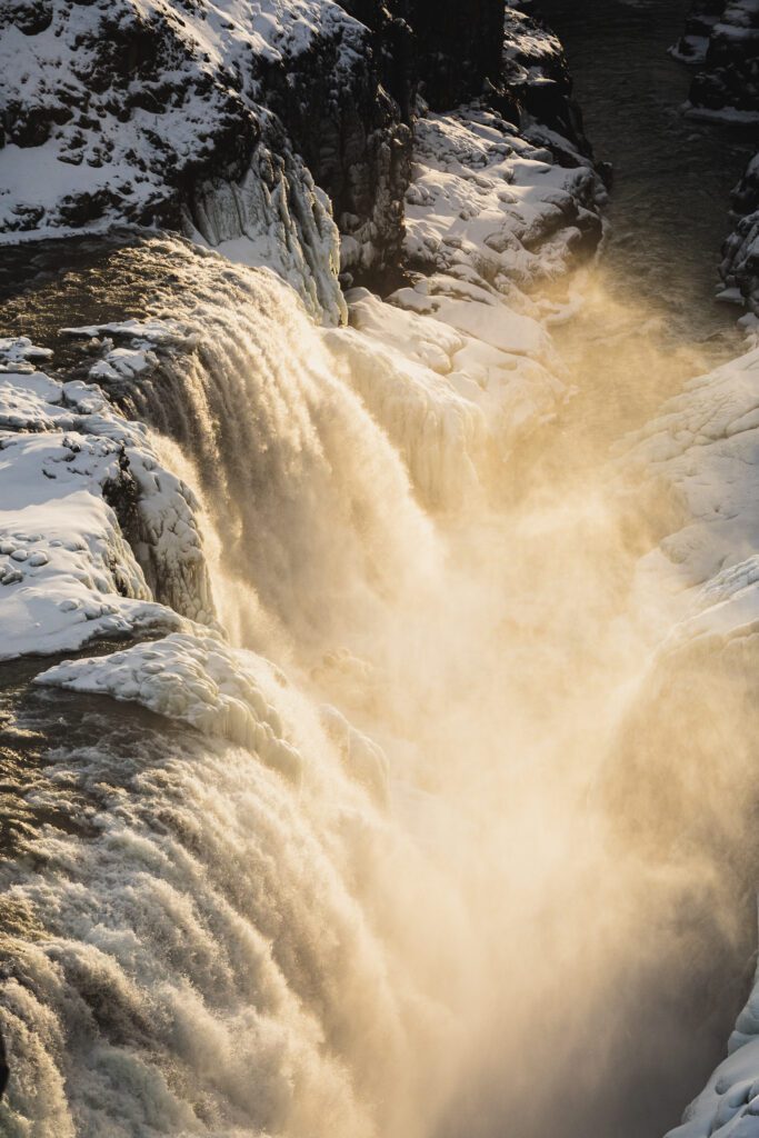 Sunlit steam billowing from a geothermal hot spring channel cutting through icy black rock.