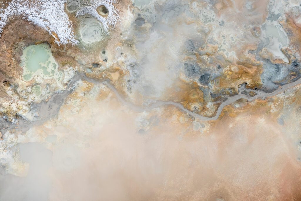 Top-down aerial of geothermal mud terraces in cyan, ochre and white mineral patterns.