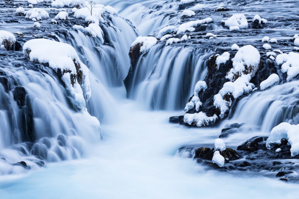 Brúarfoss in winter, vivid turquoise water cascading between snow-laden basalt steps.
