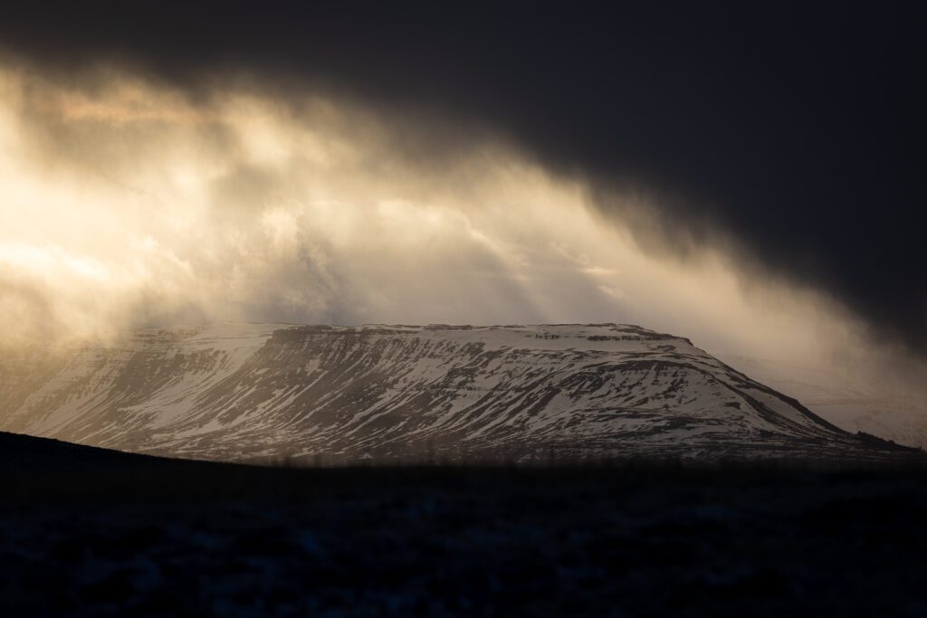 Flat-topped table mountain near Hvalfjörður catching low sun through drifting snow curtains.