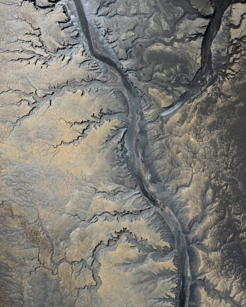 Top-down aerial of a maroon crater scarred by snow patches with a meandering blue stream below it.