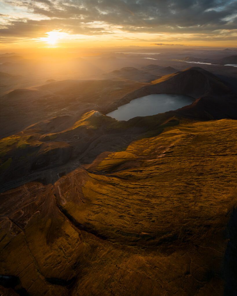Sunset aerial of a small crater lake on a high mossy plateau with a hazy basin opening beyond.