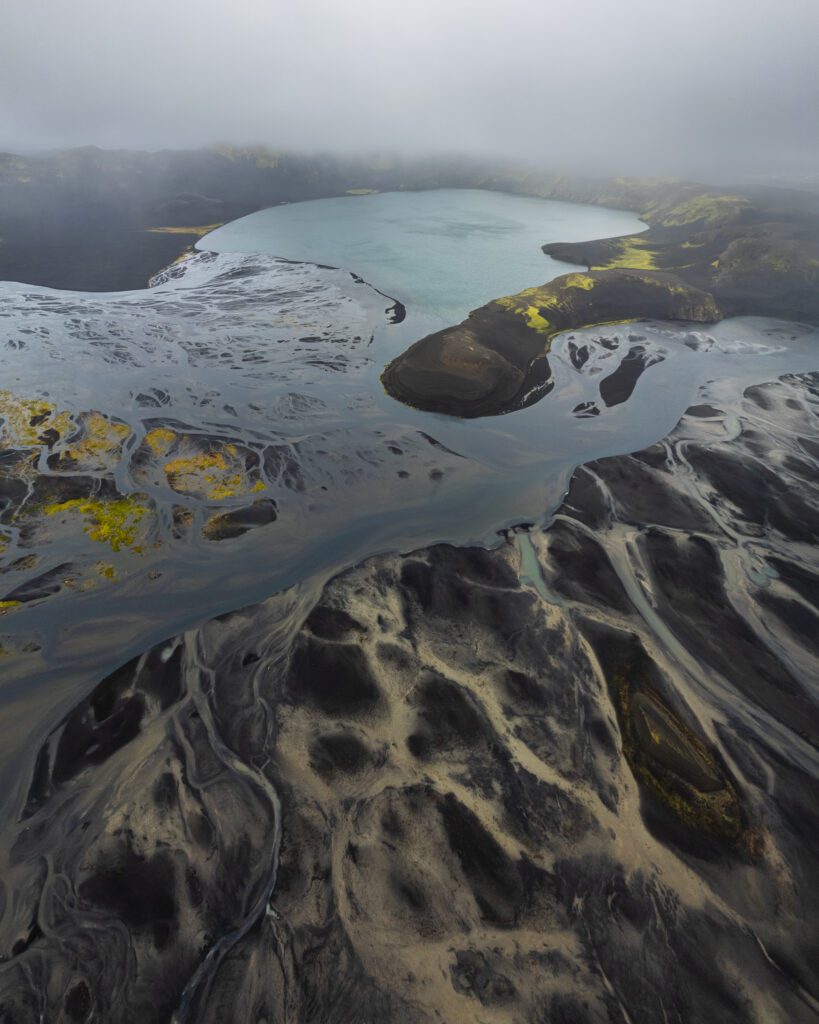 Top-down aerial of a red crater holding snow patches, with a turquoise meltwater stream curling out below.