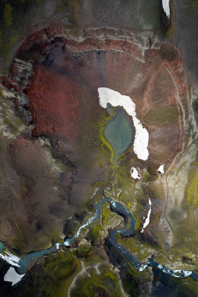 Aerial of Ljótipollur, the red-walled crater lake with deep teal water, distant mountains under heavy cloud.