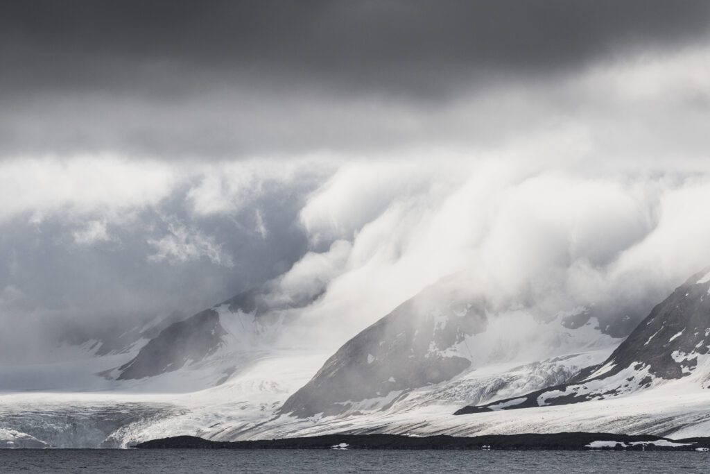 Cloud cascading down snow-covered Svalbard peaks over a glacier front.
