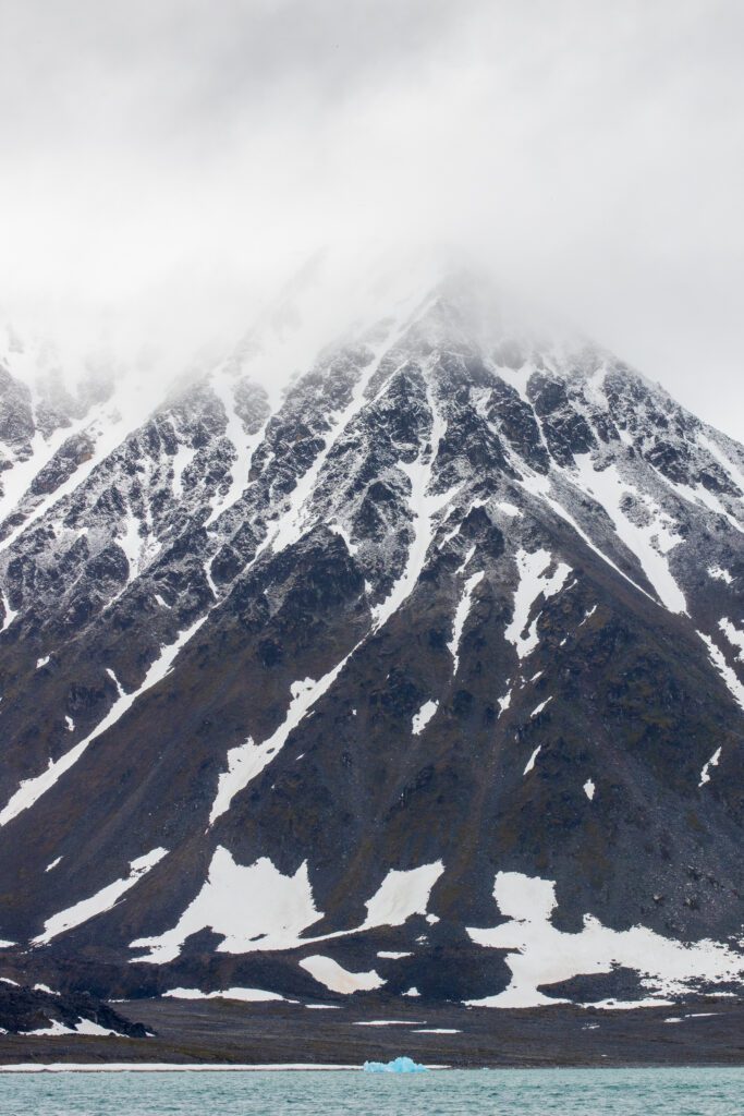Snow-streaked Svalbard peak rising from the sea with a small blue iceberg below.