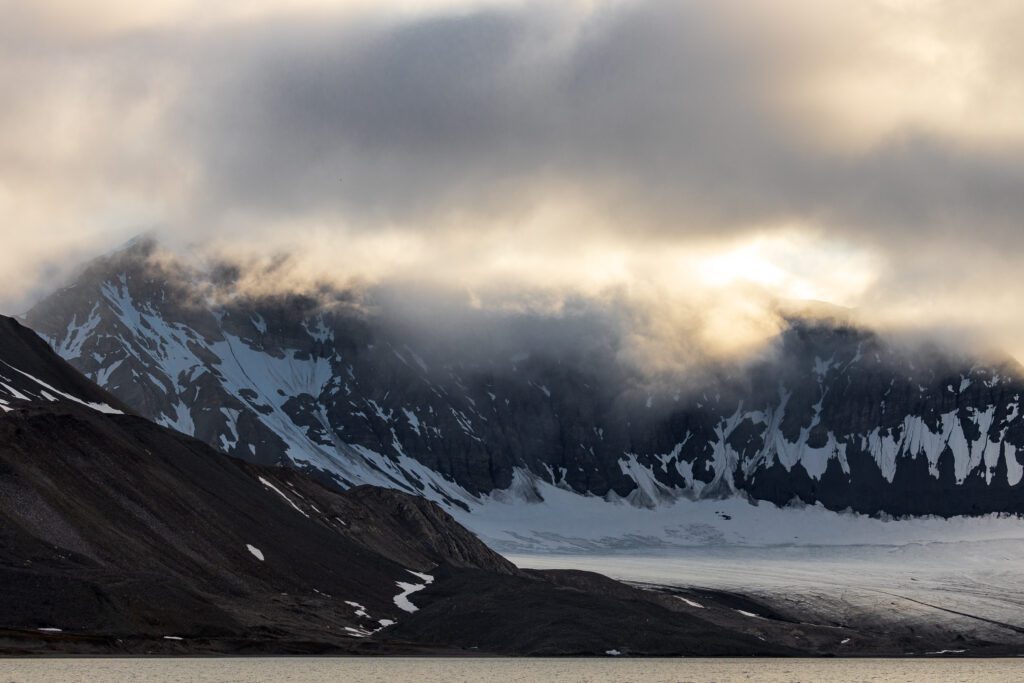 Glacier flowing between dark Svalbard peaks under low cloud at sunrise.