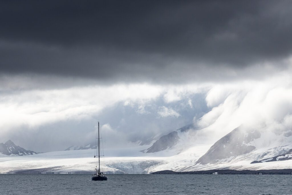 Sailboat dwarfed by Svalbard glaciers and snow-covered peaks under dark cloud.