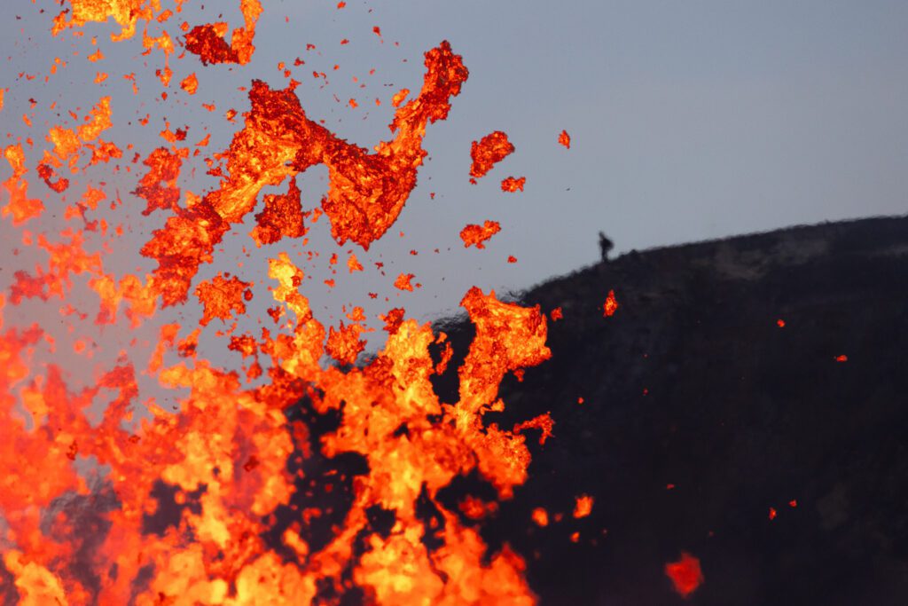 Lava bombs exploding from a fissure with a tiny silhouetted person on the ridge for scale.