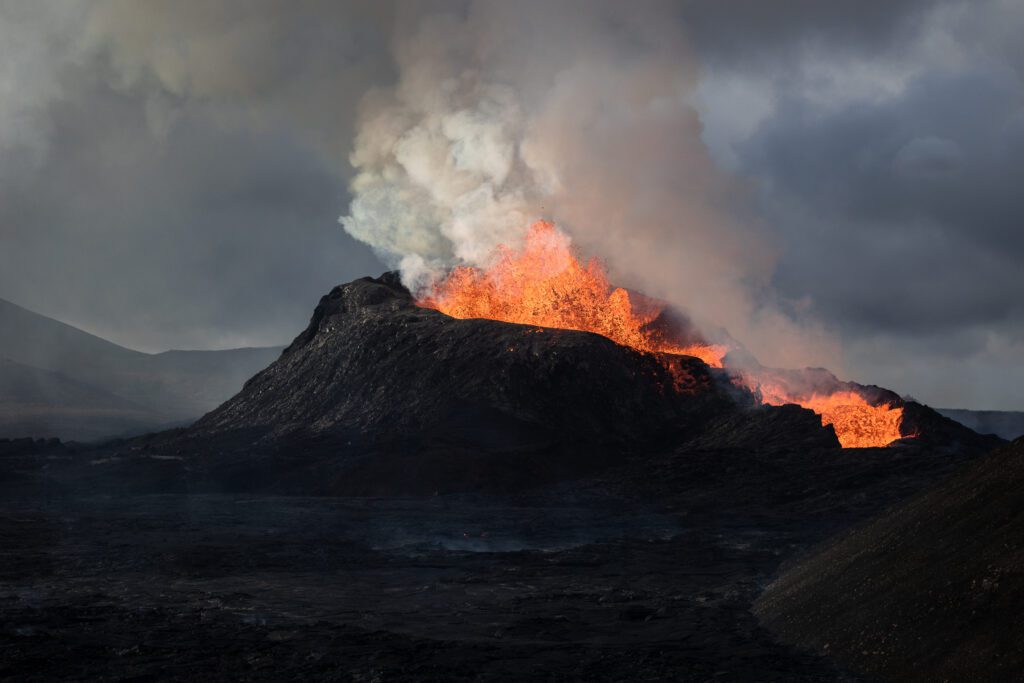Daytime cinder cone eruption with lava overflowing the rim and a smoke plume.