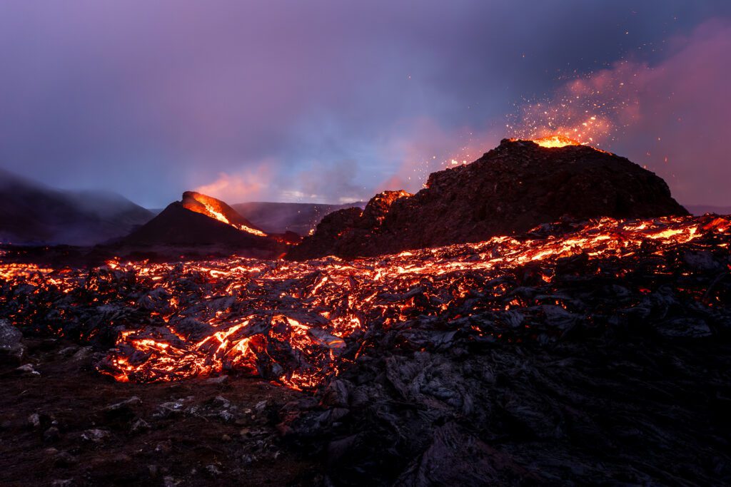Two erupting spatter cones above a glowing lava field at blue-hour dusk.