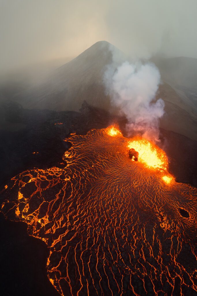 Drone view of a glowing lava vent with smoke plume against a hazy mountain backdrop.