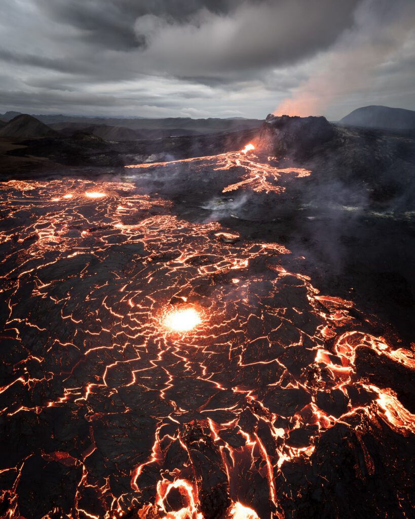 Wide aerial of a vast lava field with cracking crust and a distant cone under storm clouds.