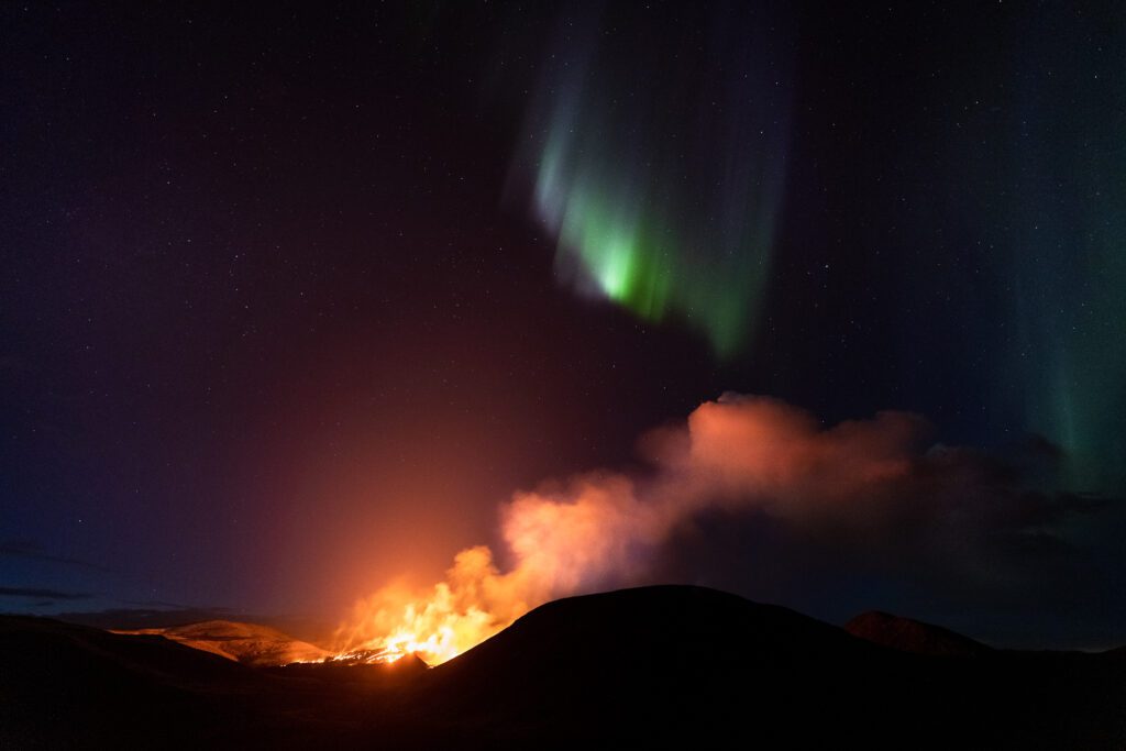 Green aurora streaking above a glowing volcanic eruption in Iceland.