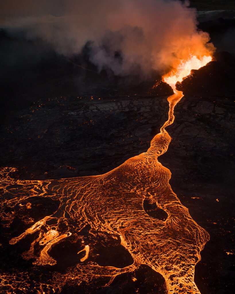 Aerial drone shot of a glowing lava river curving from a distant lava fountain.
