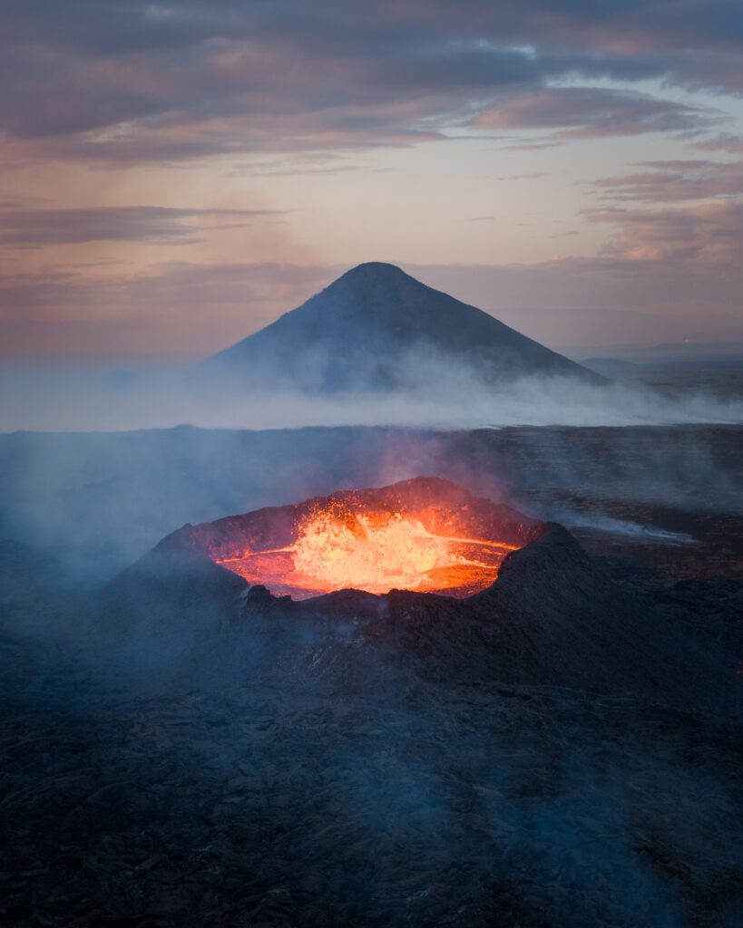 Wide aerial of an erupting crater with a bubbling lava lake at sunset.