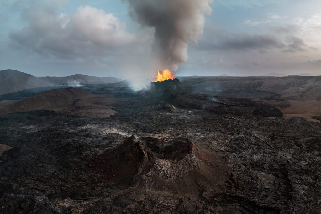Wide aerial of an erupting cinder cone with a tall smoke plume and surrounding lava field.