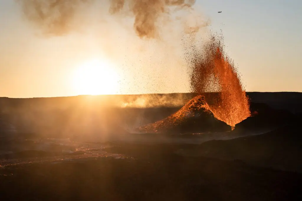 Erupting cinder cone with a lava fountain at sunrise, helicopter dot in the sky.