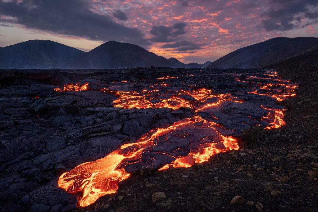 River of glowing lava through a valley at sunset with mountains behind.