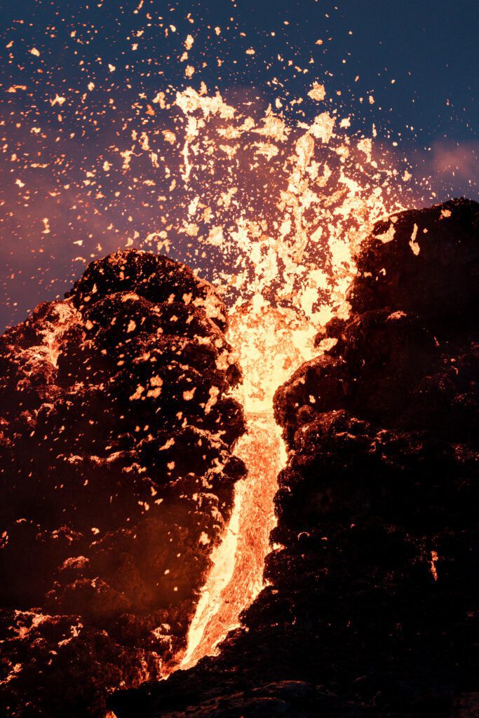 Lava fountain bursting from a cinder cone with a glowing river flowing down the slope.